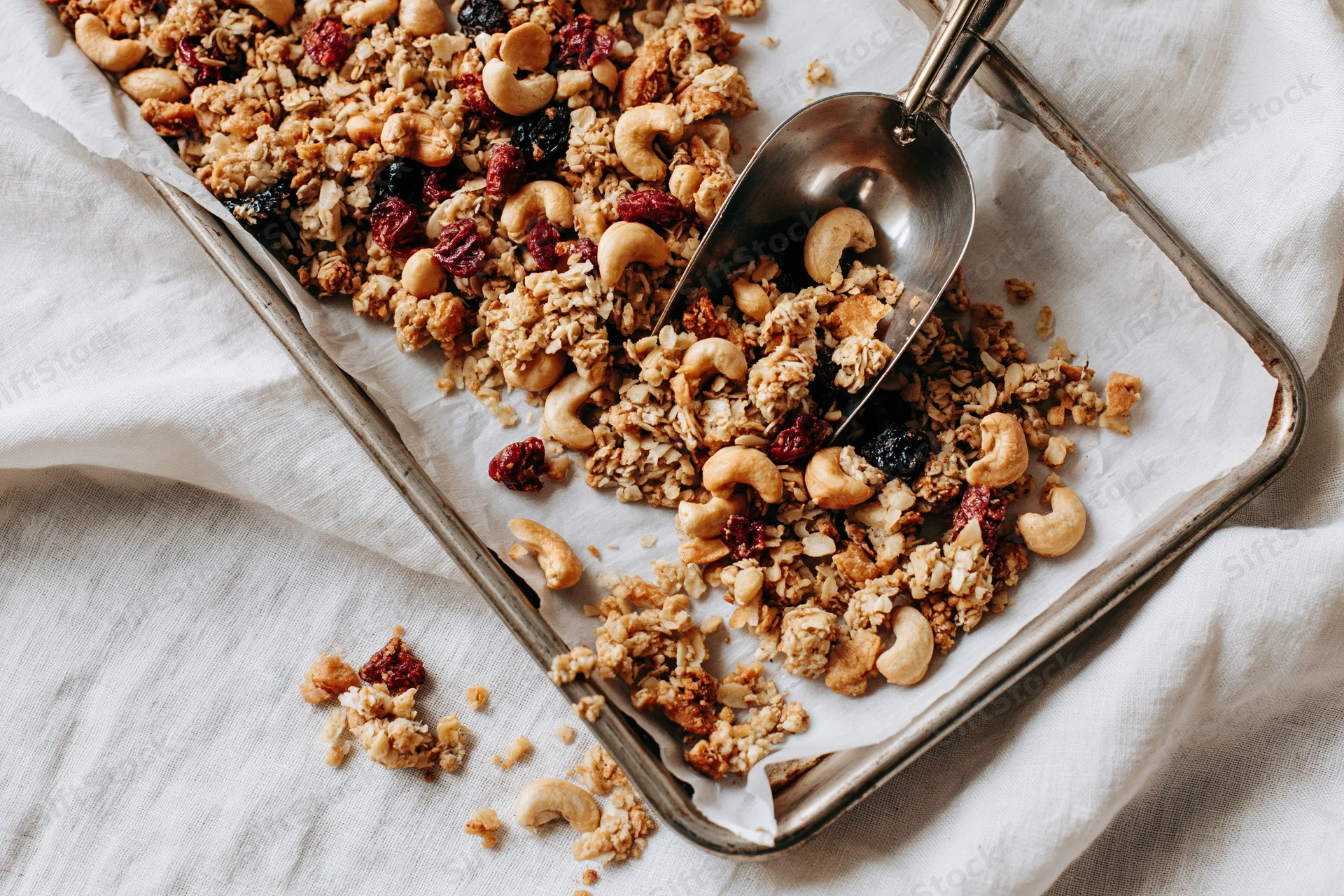 Granola with cashews and dried fruit on a parchment-lined baking tray with a metal scoop, resting on a white linen cloth