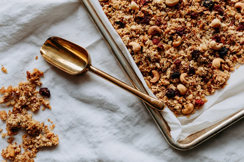 Granola with cashews and dried fruit on a baking tray with a golden spoon beside it on a white cloth