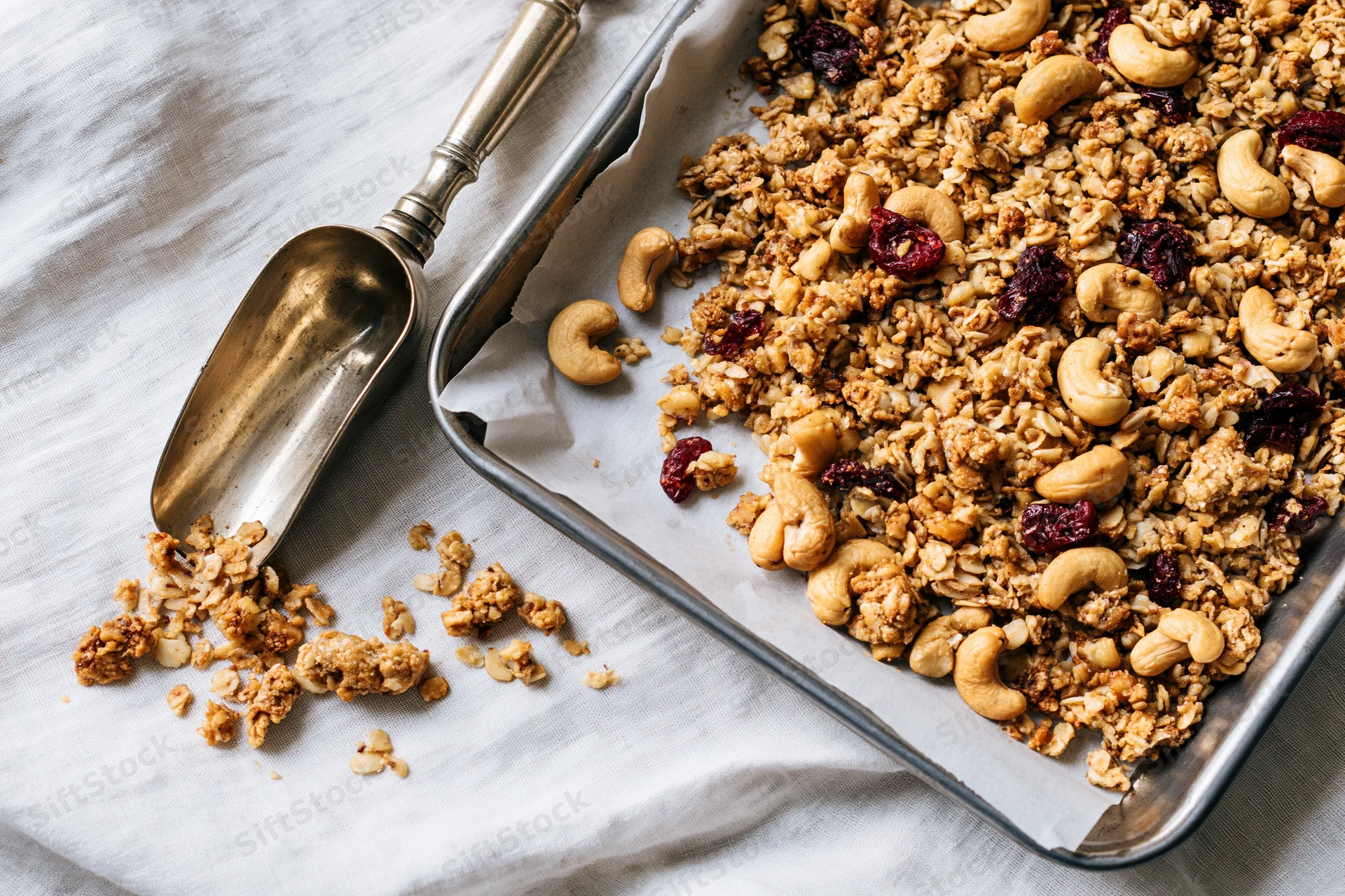 Granola with cashews and dried cranberries on a baking tray lined with parchment paper, beside a metal scoop on a cloth surface