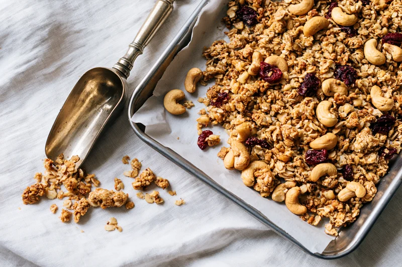 Granola with cashews and dried cranberries on a baking tray lined with parchment paper, beside a metal scoop on a cloth surface