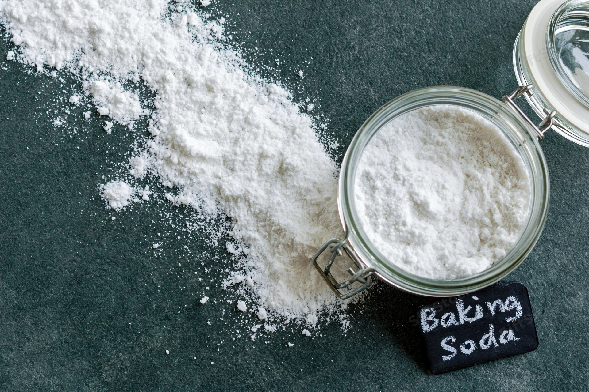 Baking soda powder spilled beside a glass jar with a chalkboard label on a dark countertop