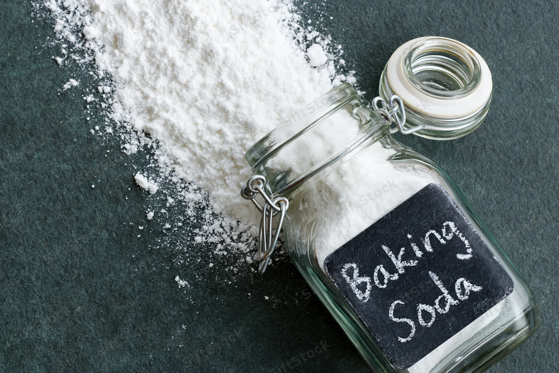 Glass jar labeled baking soda with spilled white powder on dark countertop