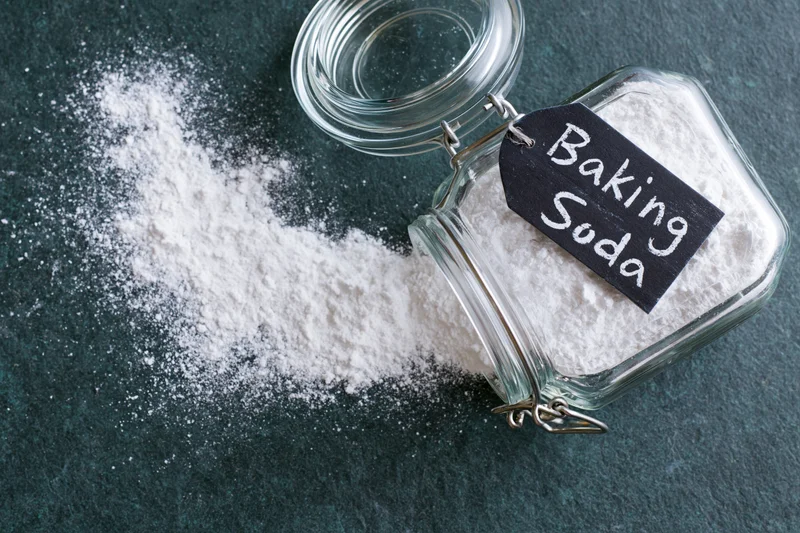 Glass jar labeled baking soda tipped over with white powder spilled onto dark countertop