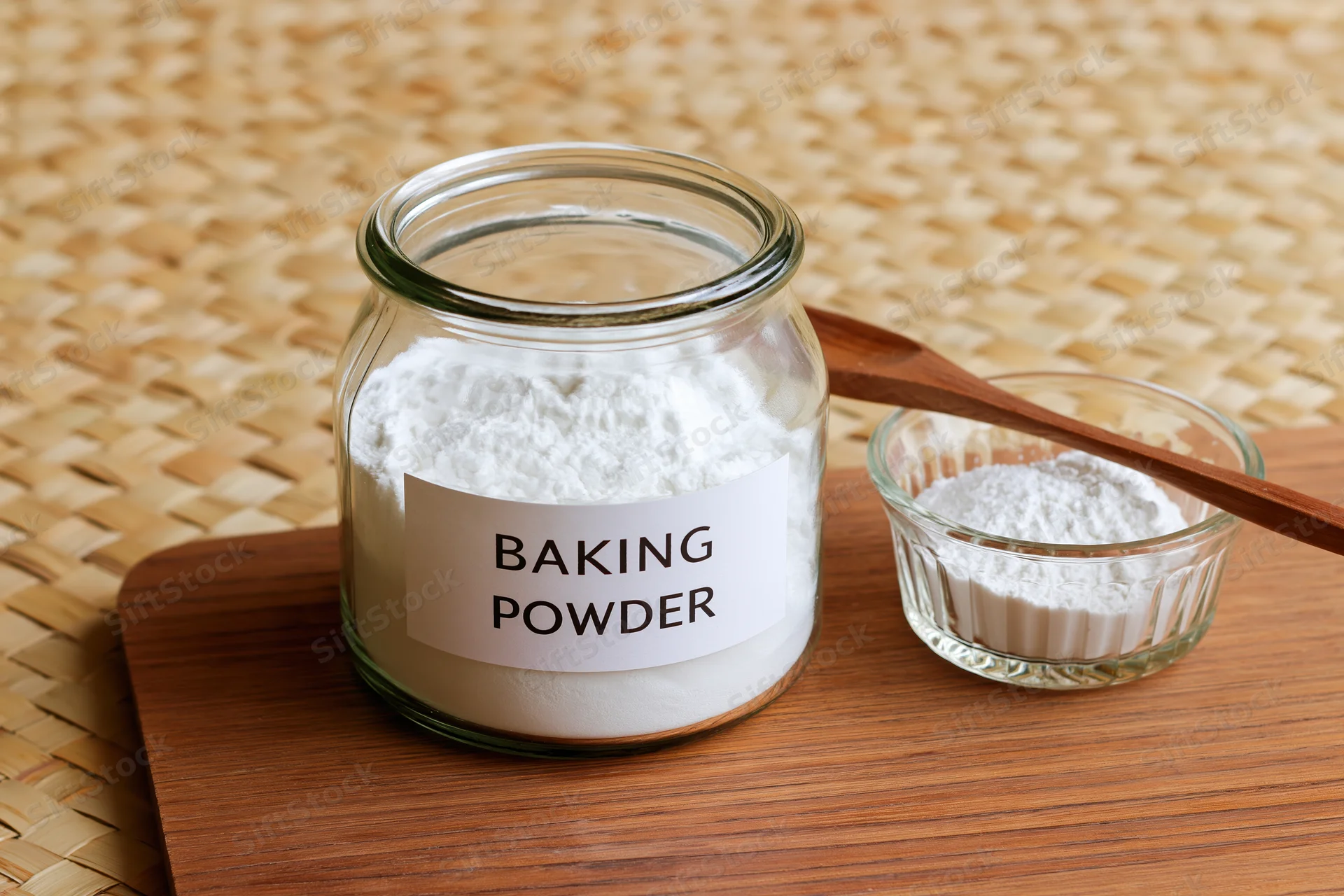 Baking powder in a labeled glass jar with a small bowl and wooden spoon on a wooden cutting board
