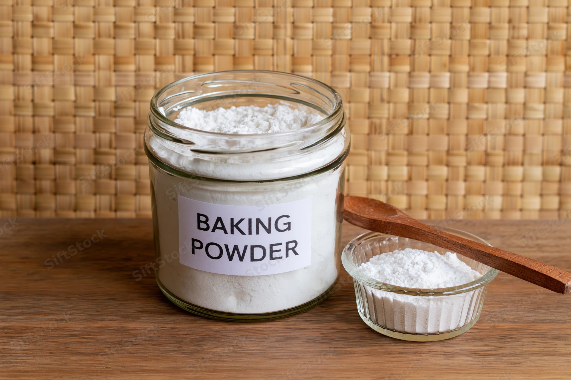 Glass jar labeled baking powder with small bowl and wooden spoon on wooden table against woven background