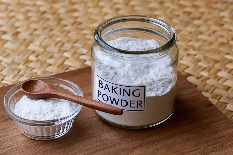 Baking powder in a glass jar with a wooden spoon on a small bowl, on a wooden table over a woven mat