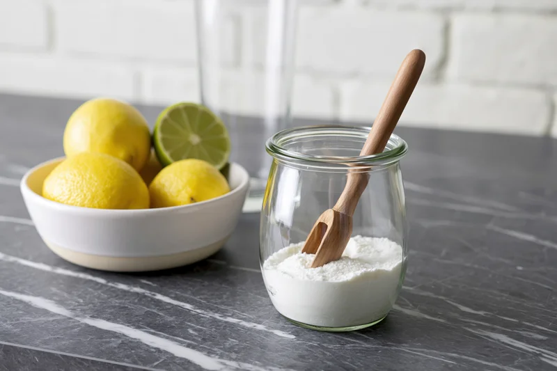Lemons and limes in a bowl beside a glass jar of flour with a wooden spoon on a marble countertop