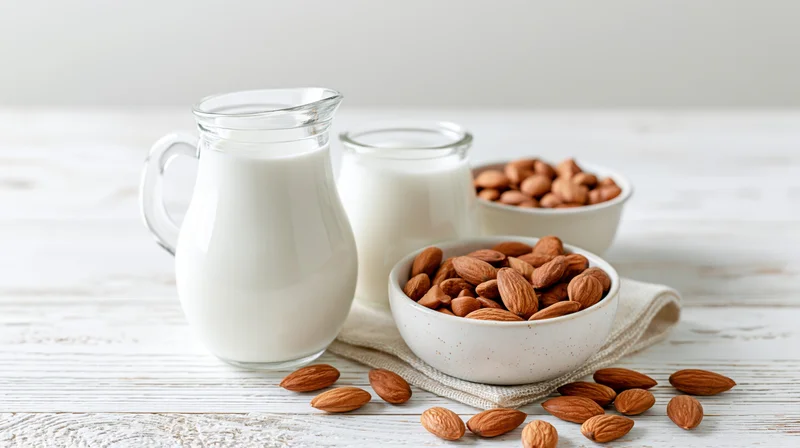 Almond milk in glass pitcher and jar beside bowls of almonds on white wooden table