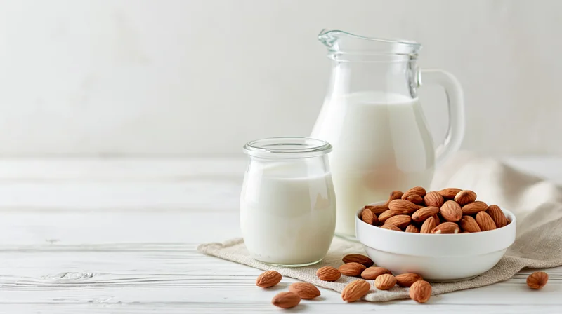 Almond milk in glass pitcher and jar beside bowl of almonds on white wooden table