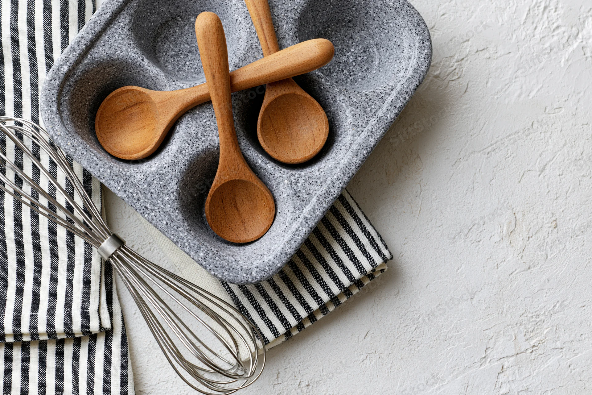 Wooden spoons and a whisk arranged in a gray dish on a striped cloth over a white textured surface