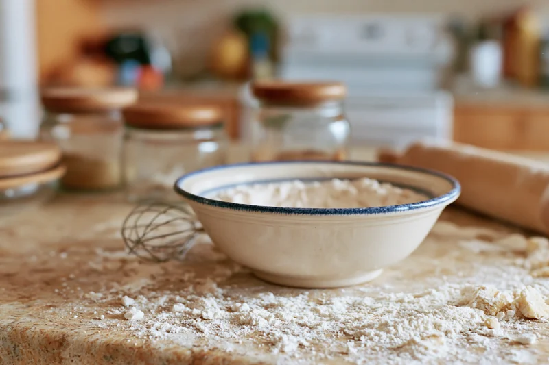 Bowl of flour on a floured countertop in a kitchen with baking tools and jars nearby