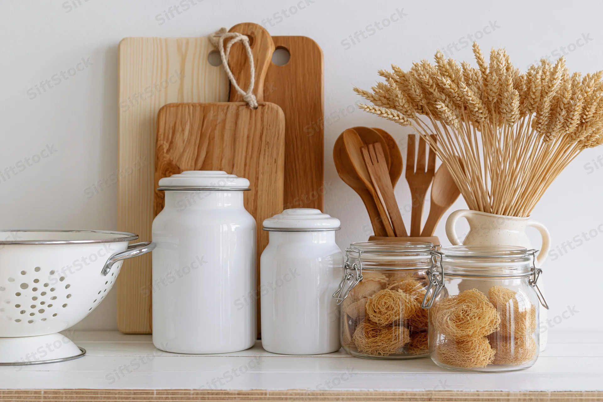 Kitchen countertop with wooden cutting boards, white enamel jars, glass jars holding dried noodles, and wheat stalks in a ceramic vase