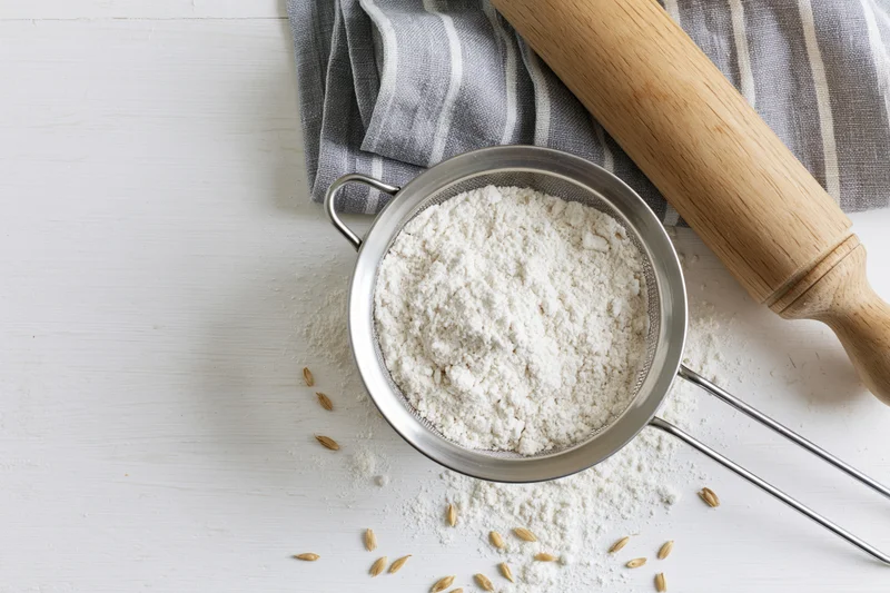Flour in a metal sifter on a white table with a wooden rolling pin and striped cloth nearby