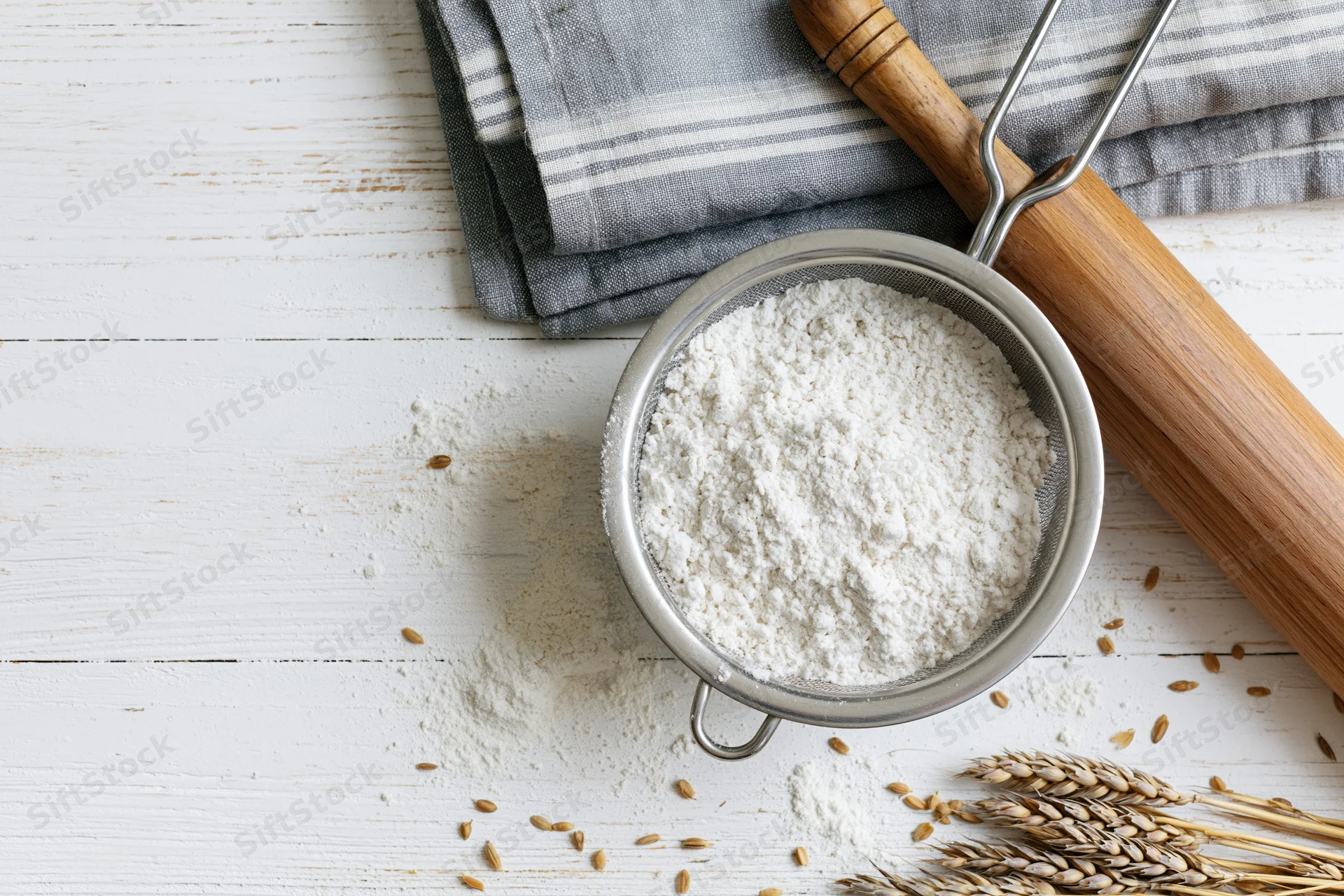 Flour in a metal sifter on a white wooden table with a rolling pin and wheat sheaf nearby
