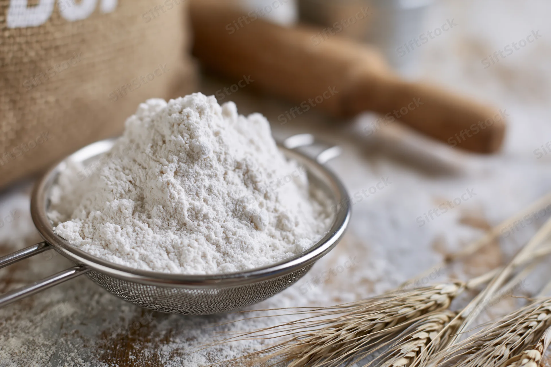 Flour in a metal sifter on a kitchen countertop with wheat stalks and a rolling pin nearby