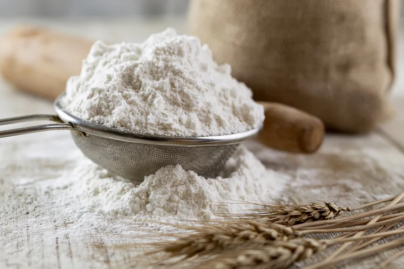 Flour in a metal sifter on a wooden surface with wheat stalks and a rolling pin nearby
