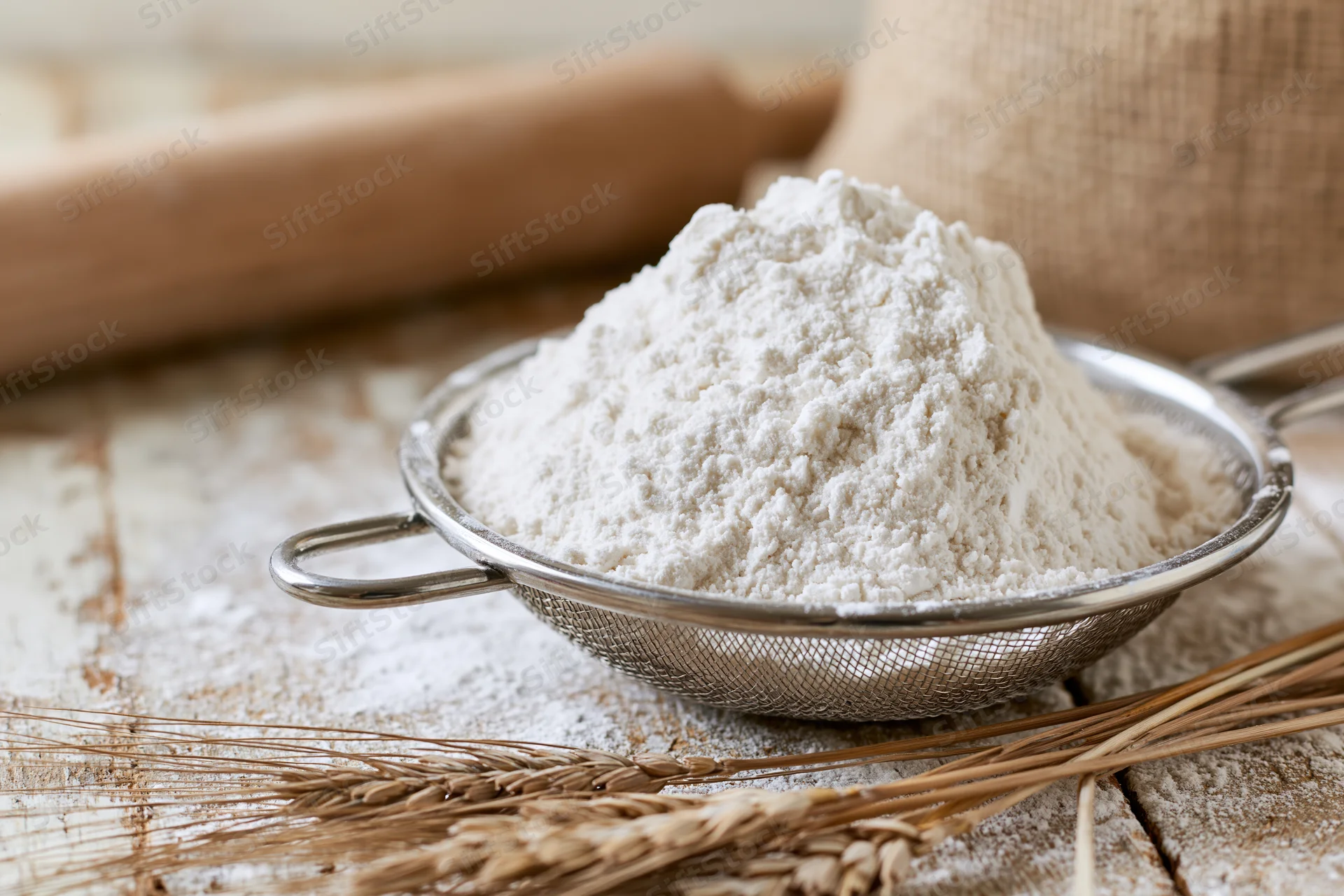Flour piled in a metal sifter on a wooden table with wheat stalks and a rolling pin nearby