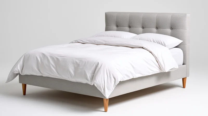 Upholstered bed with white bedding and wooden legs on a plain background