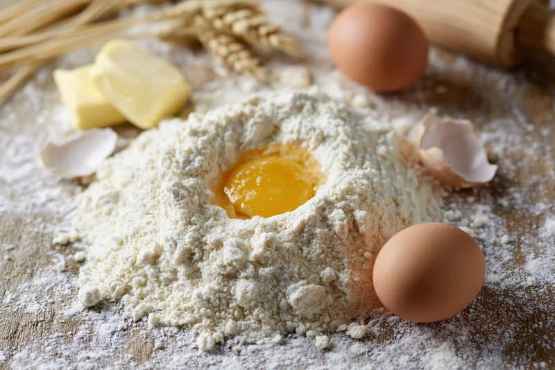 Flour mound with cracked egg in center surrounded by butter, whole eggs, wheat stalks and rolling pin on wooden surface