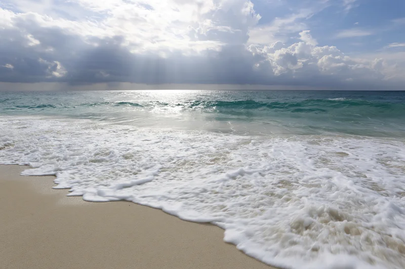 Waves breaking on sandy beach under cloudy sky with sunbeams piercing through clouds