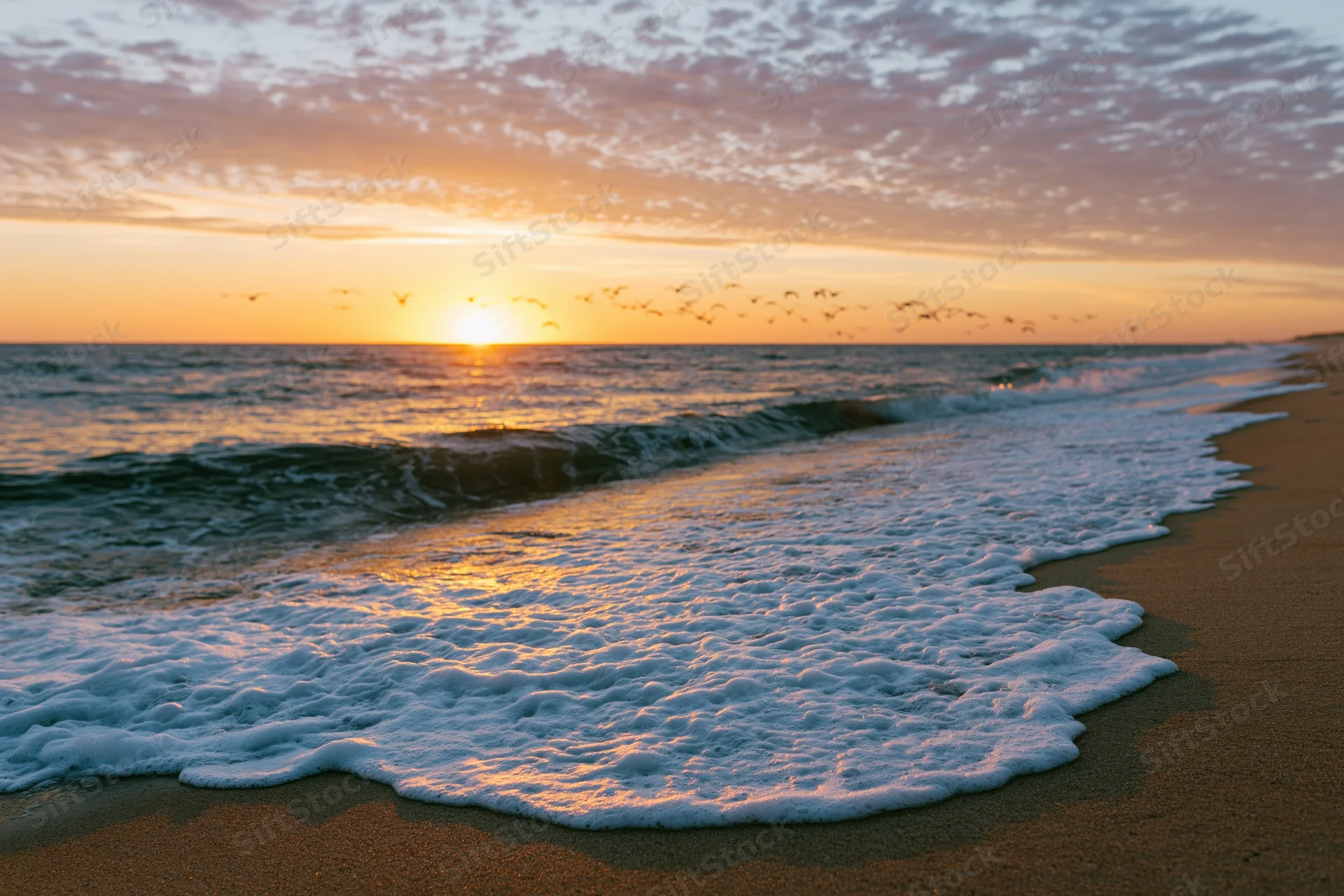 Waves with foam rolling onto sandy beach at sunset over ocean horizon