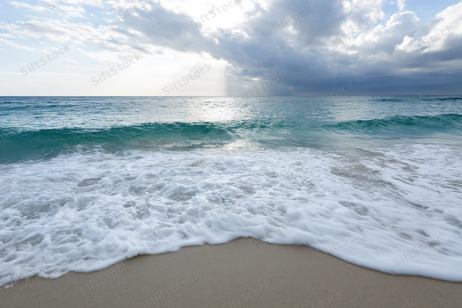 Waves breaking on sandy beach under cloudy sky with sunlight reflecting on ocean surface
