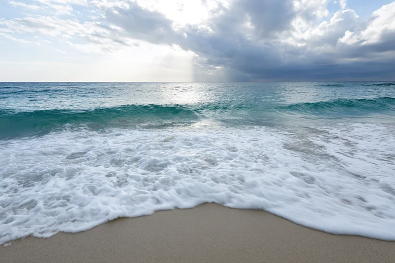 Waves breaking on sandy beach under cloudy sky with sunlight reflecting on ocean surface