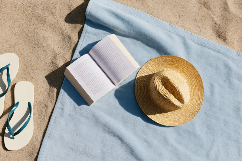 Open book and straw hat on a blue towel beside flipflops on sandy beach