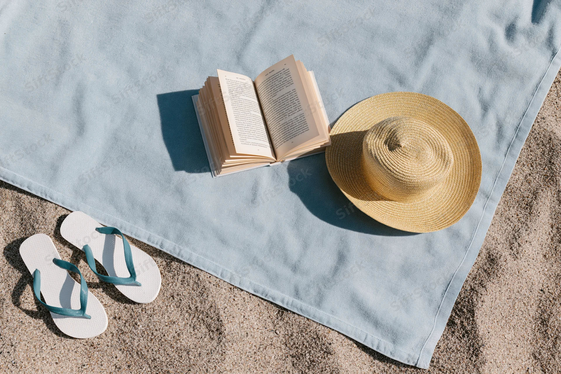 Open book and straw hat on a blue towel with flipflops on sandy beach