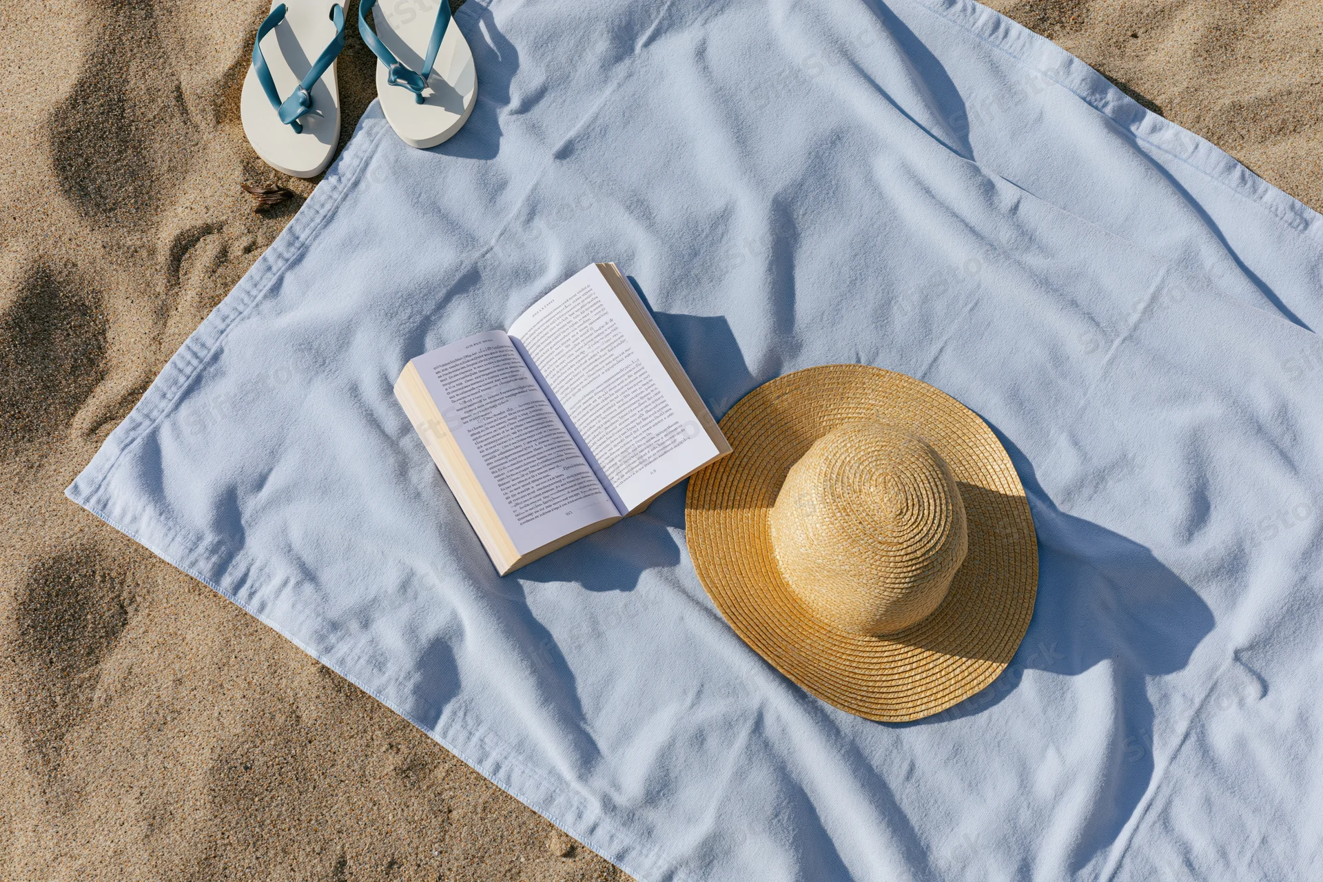 Open book and straw hat on a blue towel spread on sandy beach with flipflops nearby