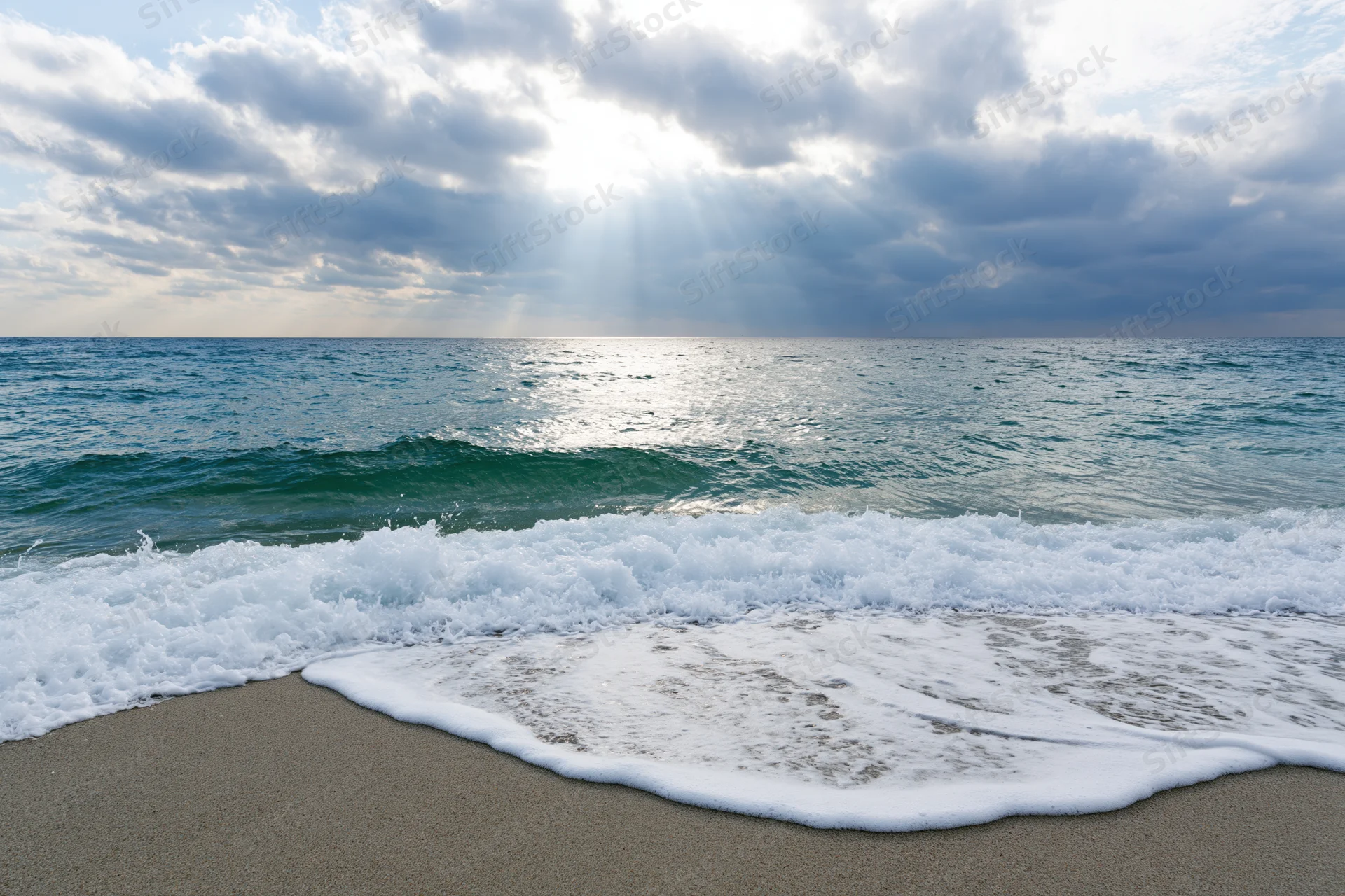 Waves breaking on sandy beach under cloudy sky with sunbeams piercing through clouds