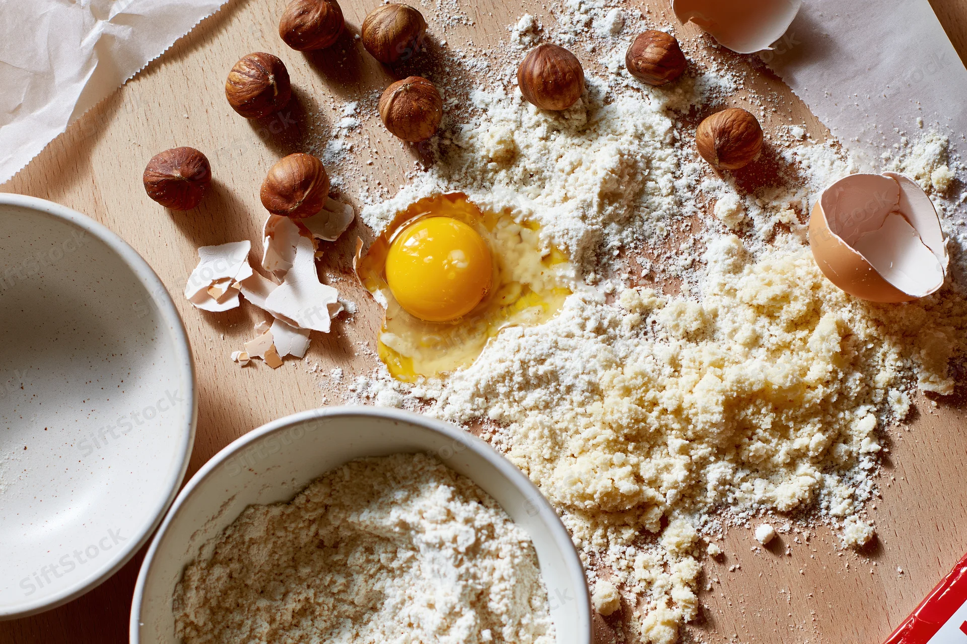 Cracked egg and scattered flour with hazelnuts on wooden countertop near bowls