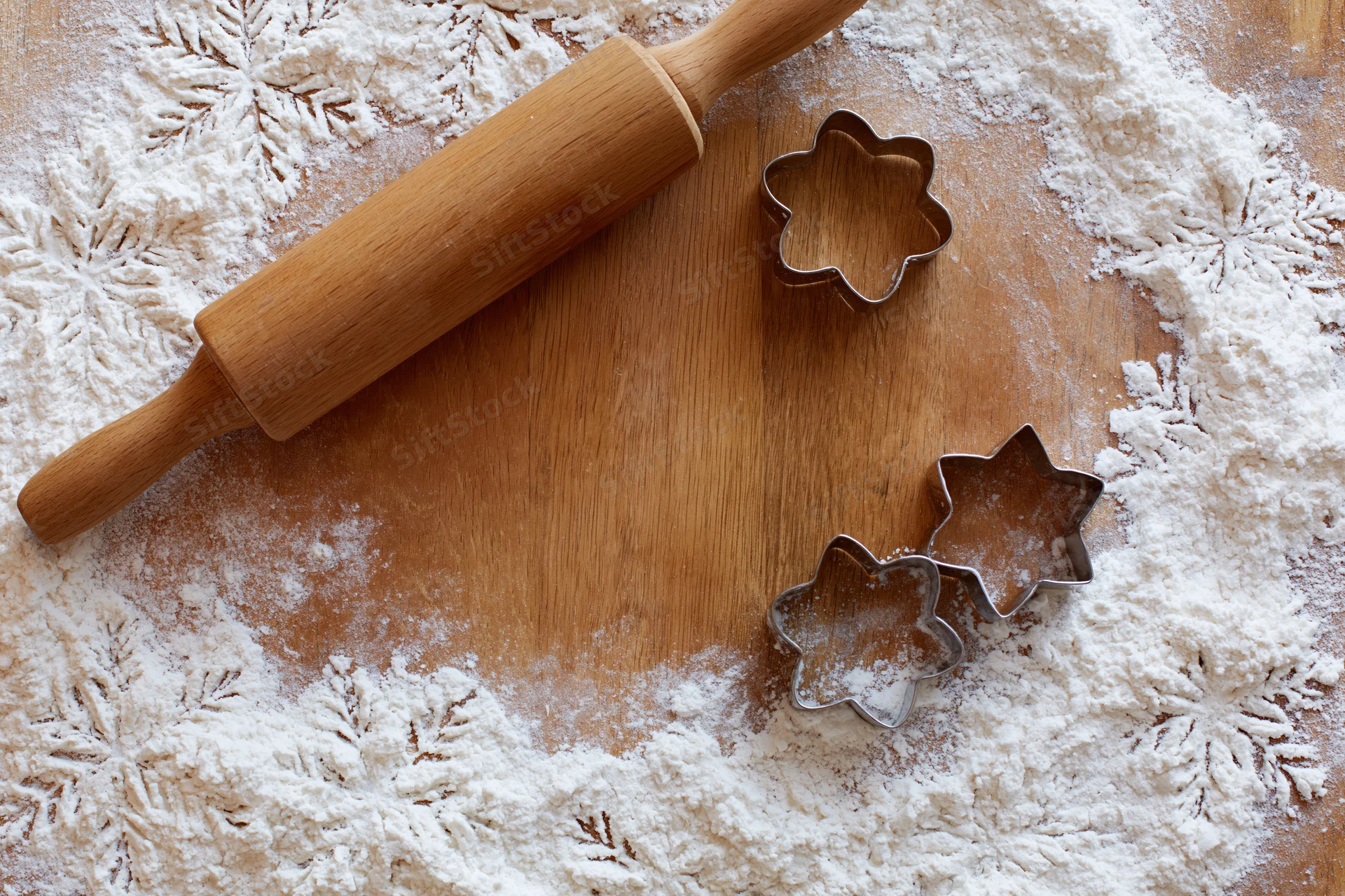 Wooden rolling pin and star-shaped cookie cutters on a floured wooden surface with snowflake patterns for holiday baking
