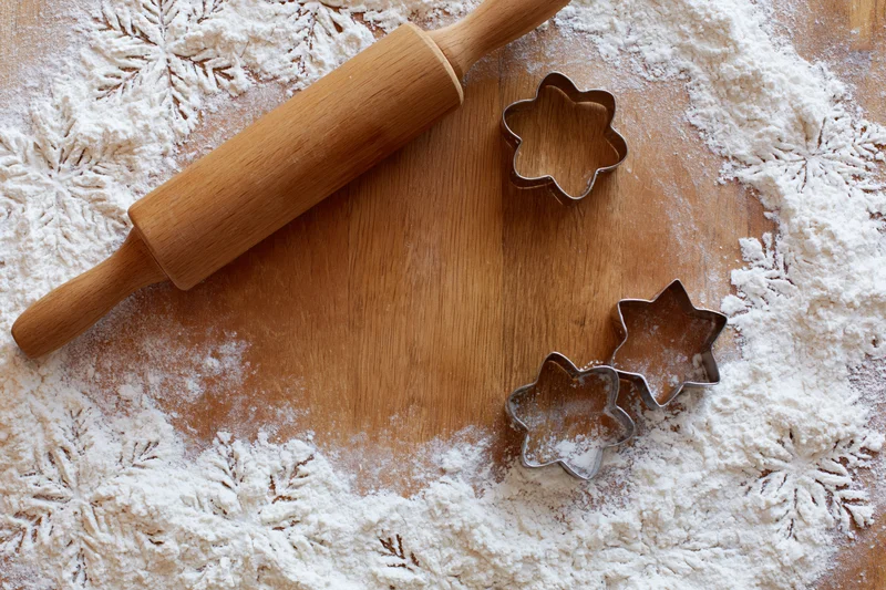 Wooden rolling pin and star-shaped cookie cutters on a floured wooden surface with snowflake patterns for holiday baking