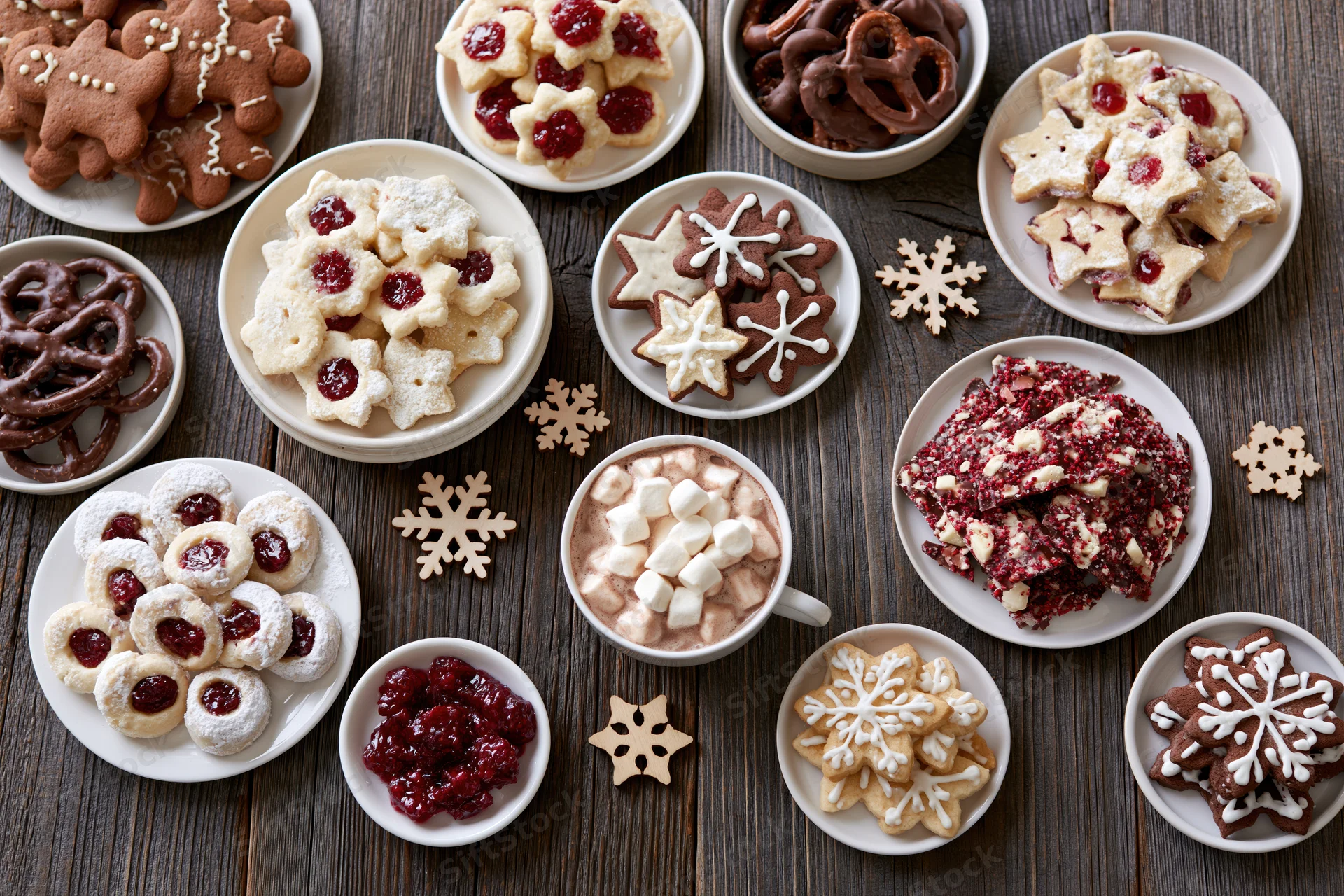 Assorted christmas cookies and desserts arranged on a wooden table with snowflake decorations and a mug of hot chocolate with marshmallows