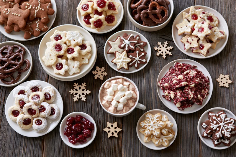 Assorted christmas cookies and desserts arranged on a wooden table with snowflake decorations and a mug of hot chocolate with marshmallows