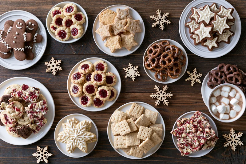 Assorted christmas cookies and sweets arranged on white plates on a dark wooden table with snowflake decorations