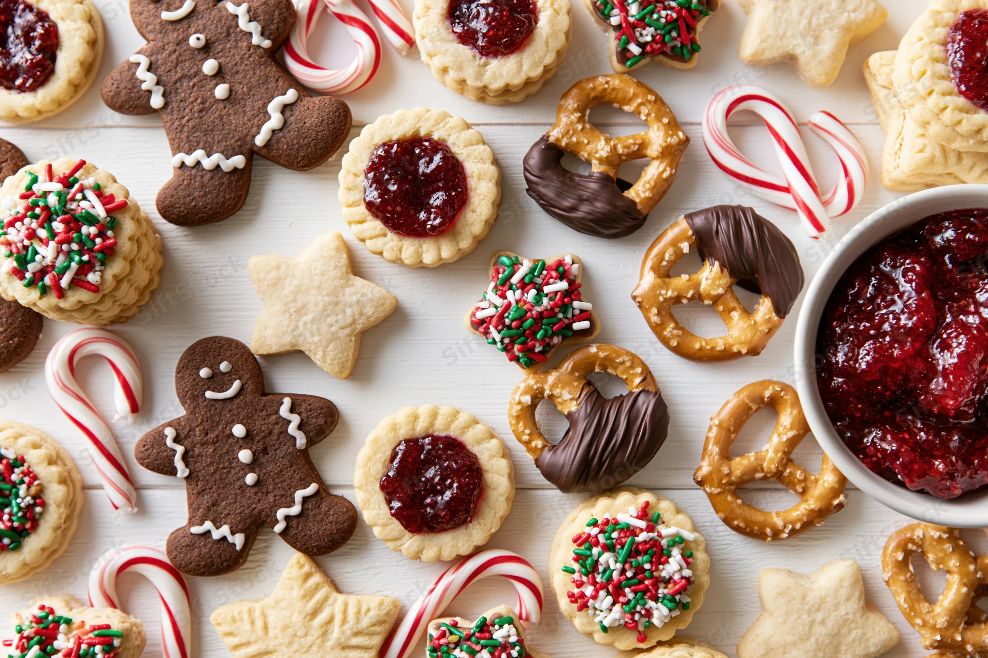 Assorted christmas cookies and candies arranged on a white wooden table with a bowl of jam and candy canes