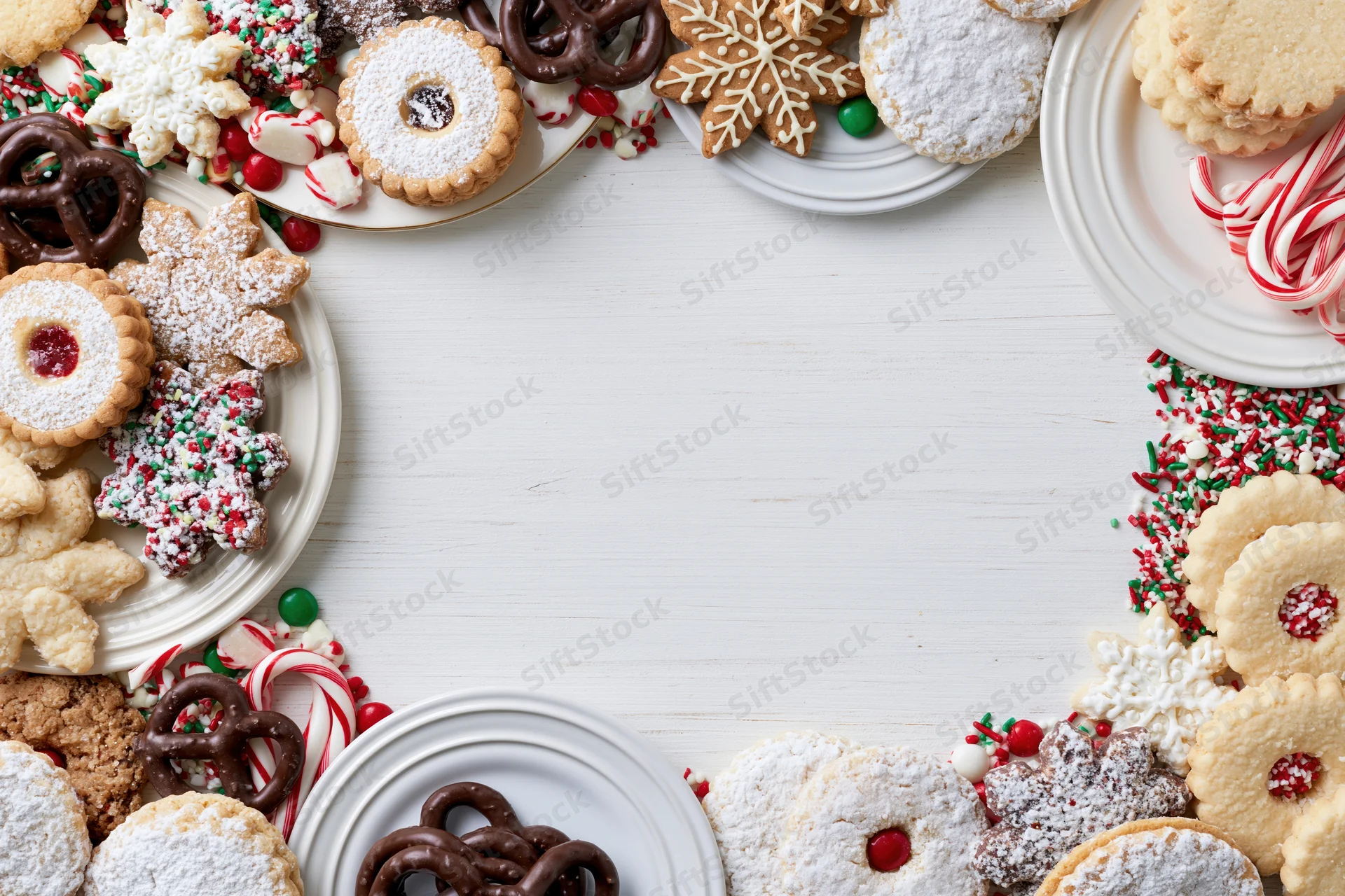 Assorted christmas cookies and candy arranged on white plates around a central copyspace on a white wooden table