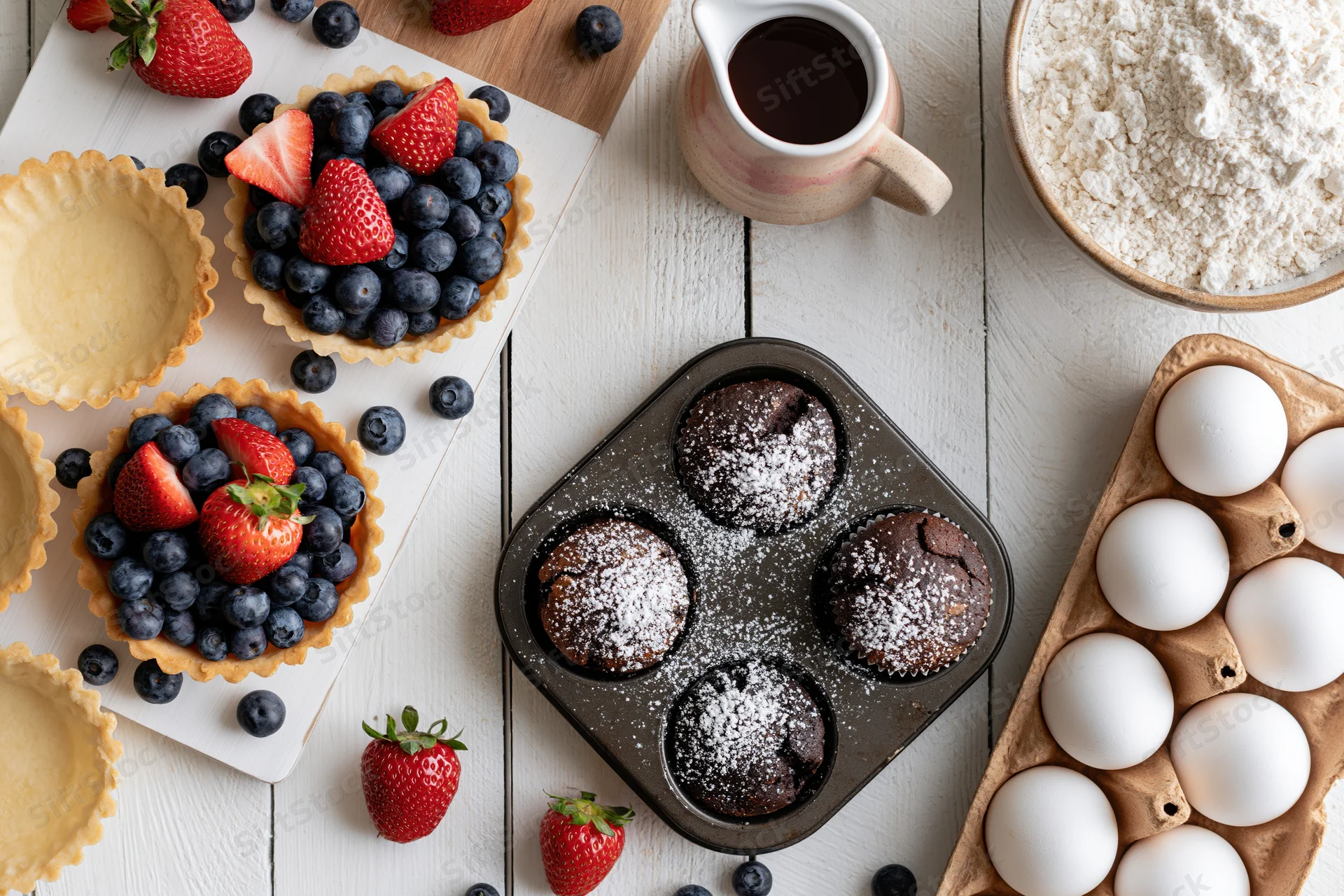 Fresh fruit tarts and chocolate muffins arranged on a white wooden table with baking ingredients around
