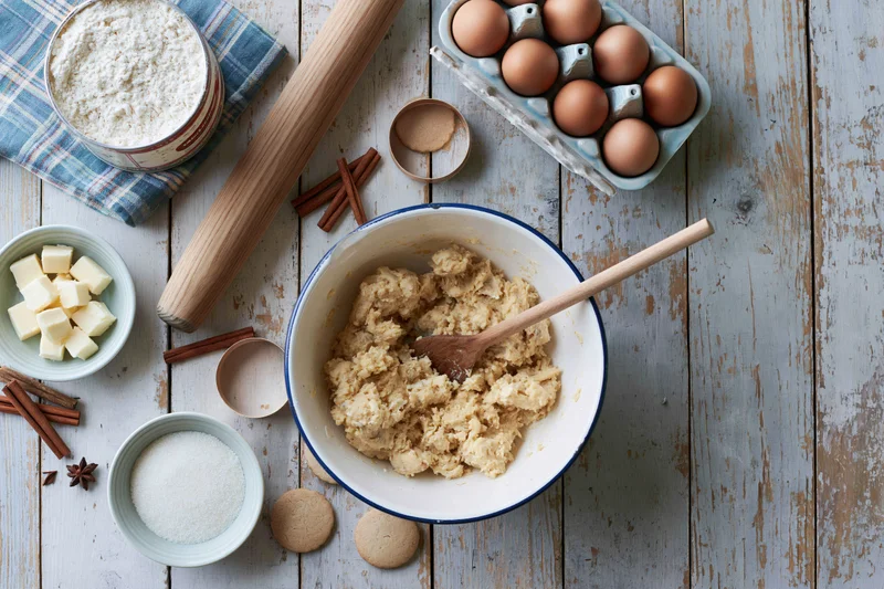 Dough mixture in a bowl with wooden spoon surrounded by baking ingredients on a rustic wooden table
