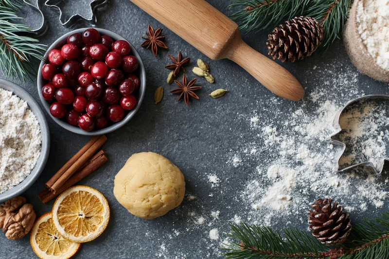 Holiday baking setup with dough, cranberries, spices and cookie cutters on a dark countertop