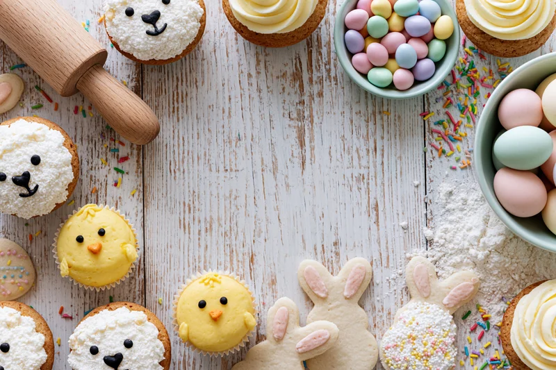 Easter-themed cupcakes and cookies shaped like bunnies and chicks arranged on a rustic white wooden table with sprinkles, flour, and a rolling pin