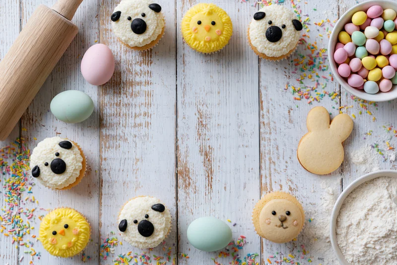 Easter cupcakes decorated with animal faces and pastel eggs on a rustic wooden table with baking ingredients