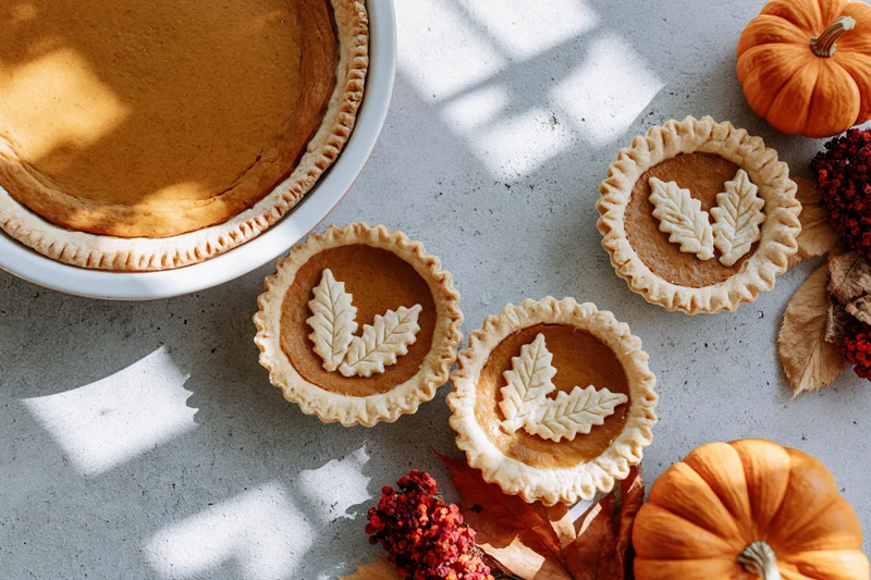 Pumpkin pie and mini tarts with leaf decorations on a countertop surrounded by pumpkins and autumn foliage