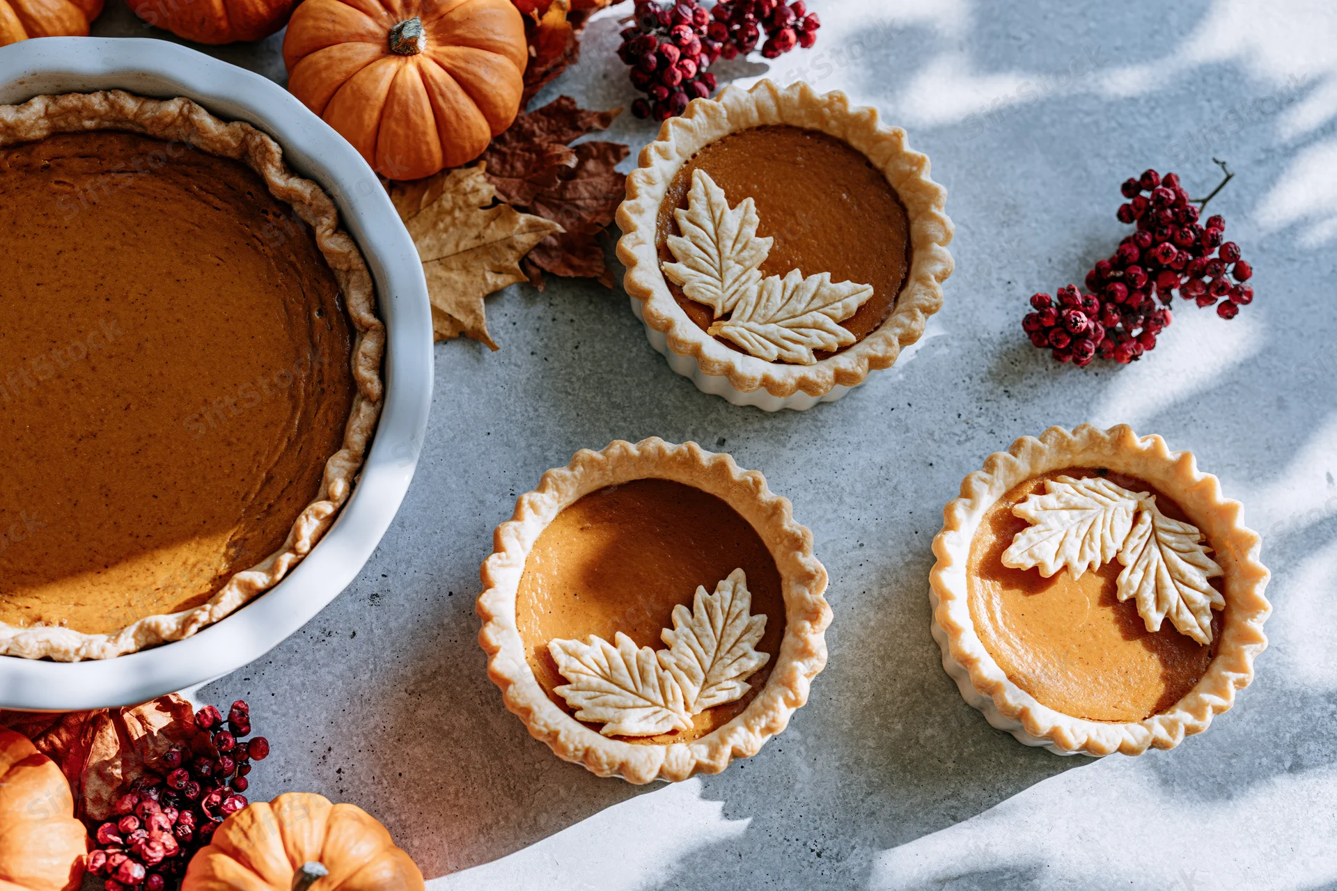 Pumpkin pie and mini tarts with leaf decorations on a table surrounded by pumpkins and berries