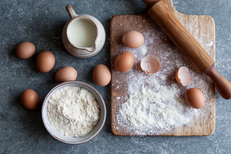 Baking ingredients arranged on a wooden cutting board and countertop with flour, eggs, milk and a rolling pin