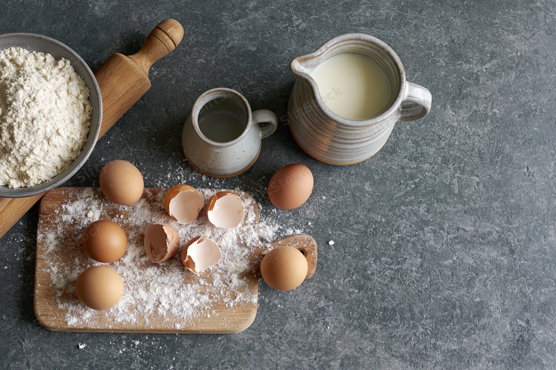 Baking ingredients including flour, eggs, milk, and eggshells arranged on a wooden cutting board and gray countertop