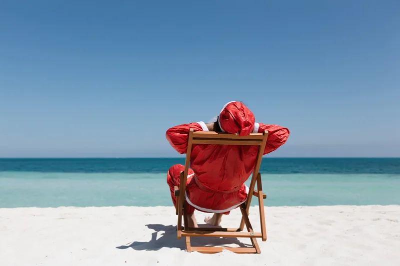 Santa reclining in a deckchair on a sandy beach facing the ocean