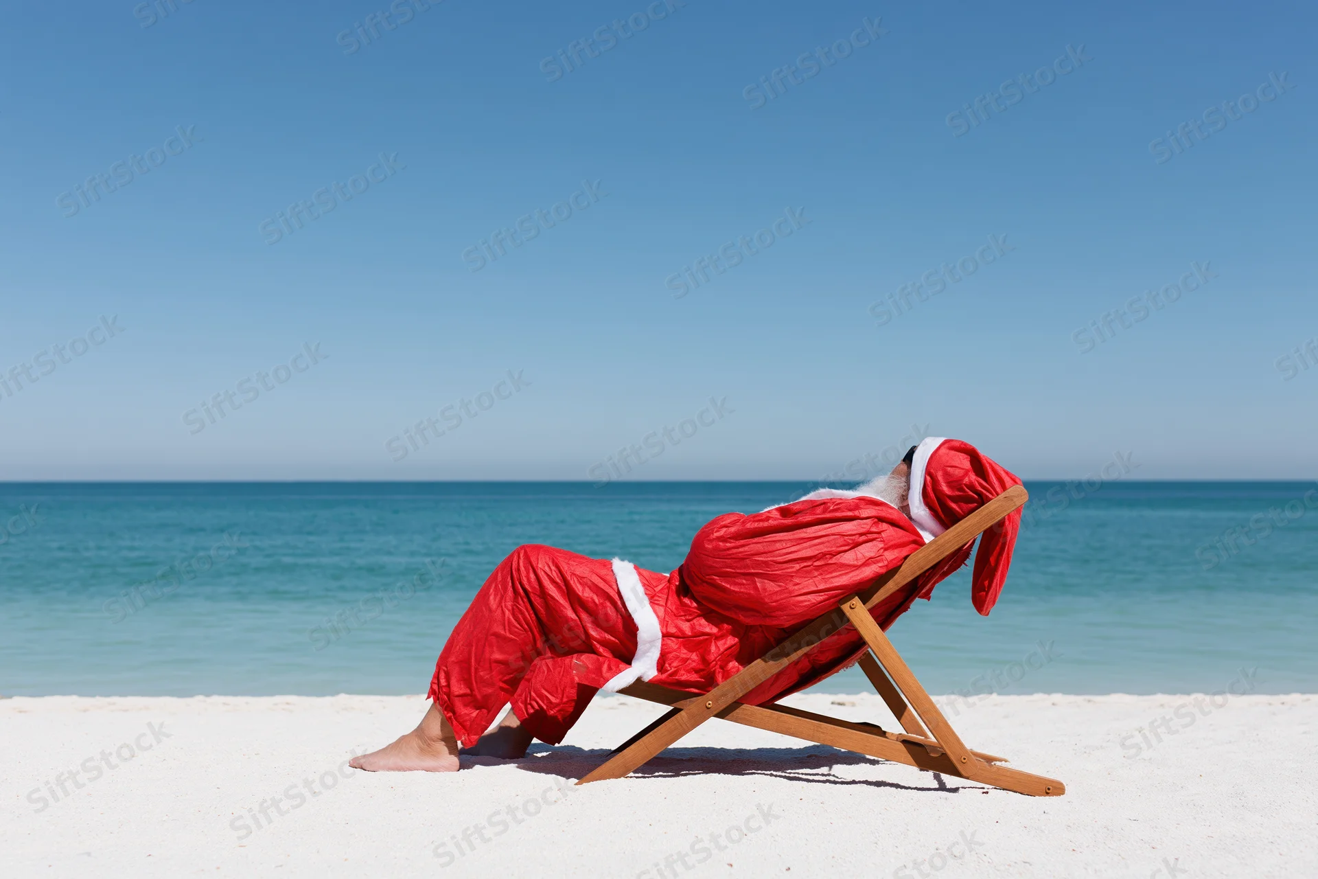 Santa reclining in a deckchair on a sandy beach beside the ocean